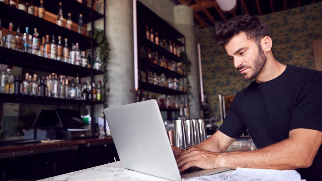 A man in a black t-shirt sits at a bar counter, surrounded by shelves of bottles, working on his laptop. Perhaps he's researching how to transfer a liquor license in California.